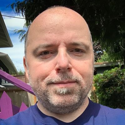 Close-up headshot of a middle-aged man with a bald head and trim beard standing in sunlight with greenery behind him.