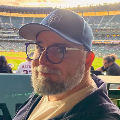 Close-up headshot of a middle-aged man in an Everton FC cap smiling at a Mariners game with the field in the background.