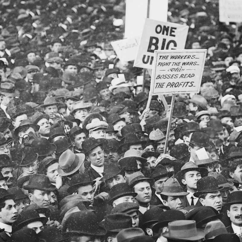 Vintage photo of a massive assembly of workers protesting with signs for the IWW ('One Big Union') and against war ('the workers fight wars … the bosses reap the profits').