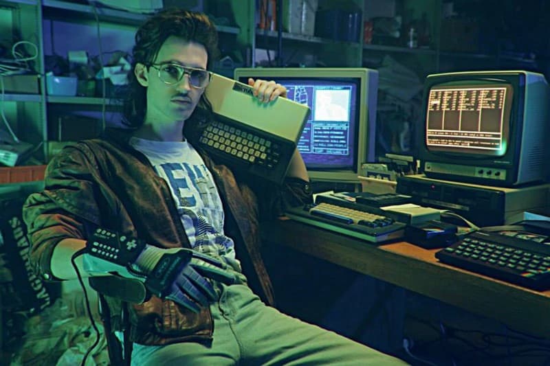 Hackerman from the short film Kung Fury sits before his computers in a leather jacket with a keyboard in one hand and a modified Nintendo Power Glove in the other.