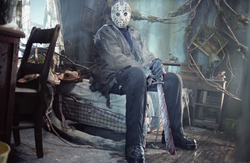 Jason from the Friday the 13th horror franchise sitting on a bed in a weed infested cabin, casually holding a macheteand looking into the camera.
