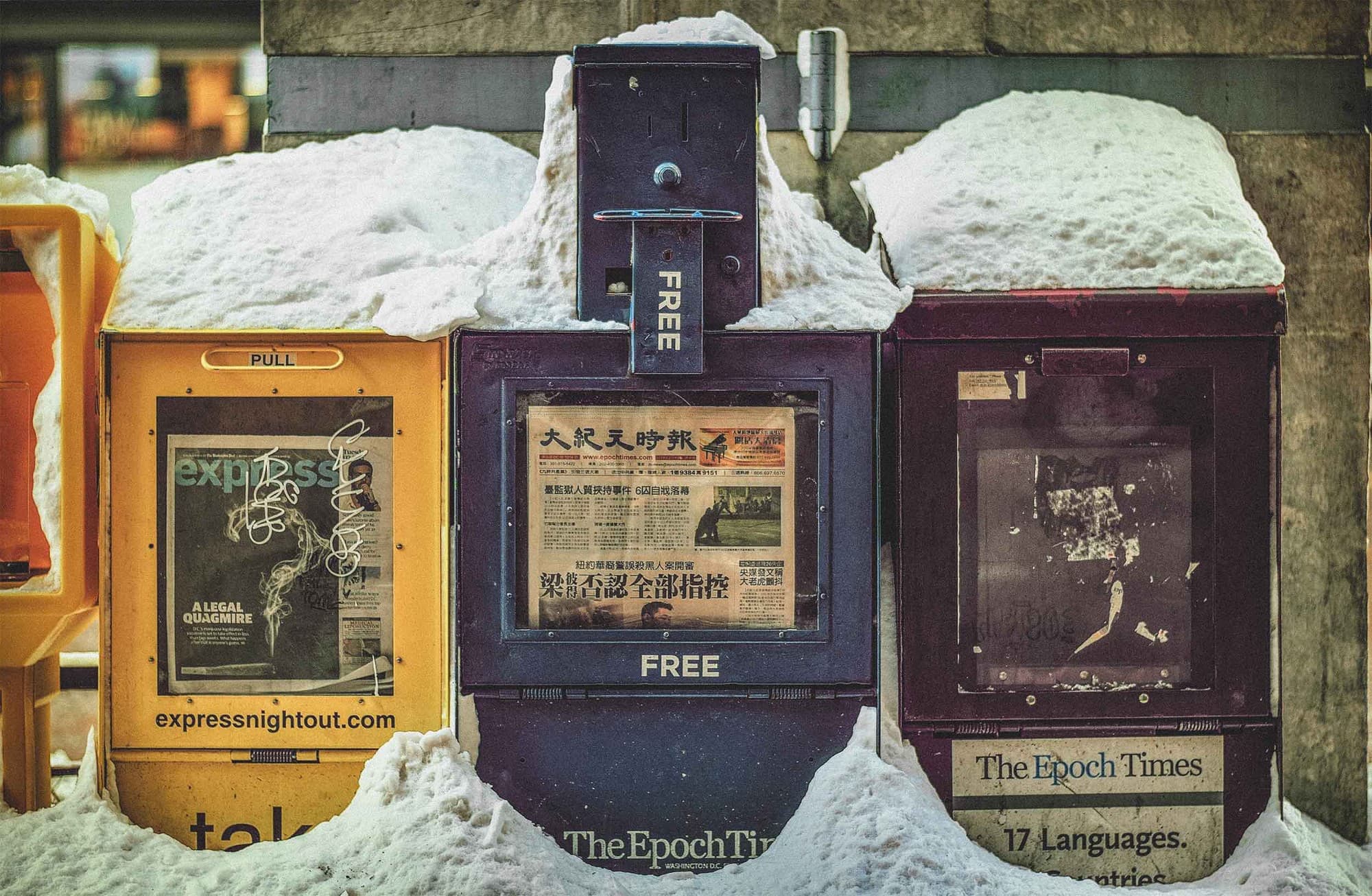 A series of newspaper vending machines in the snow.