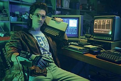 Hackerman from the short film Kung Fury sits before his computers in a leather jacket with a keyboard in one hand and a modified Nintendo Power Glove in the other.